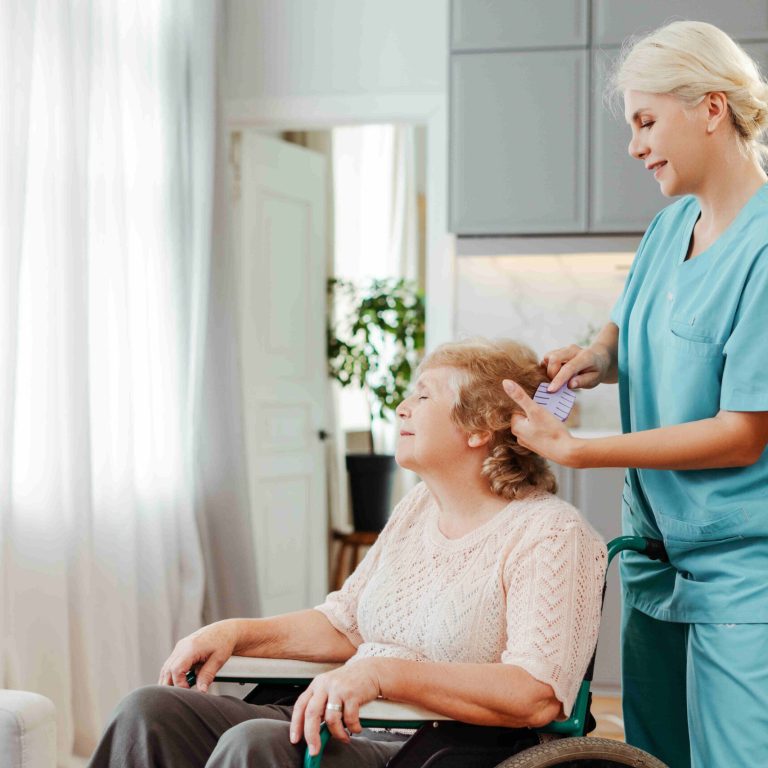 Nurse brushing hair of senior woman using wheelchair in bright living room at home. Health care concept