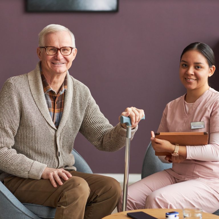 Portrait of senior man and social worker sitting on armchairs in living room and smiling at camera