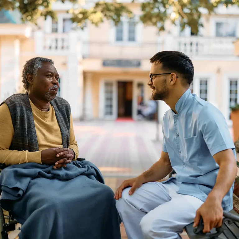 Black senior in wheelchair talking to male nurse while relaxing in the park of residential care home. Young healthcare worker talking to African American senior man who is sitting in wheelchair in the park of a nursing home.